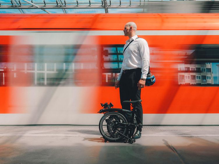 Mann mit Brompton Faltrad vor fahrendem Zug.