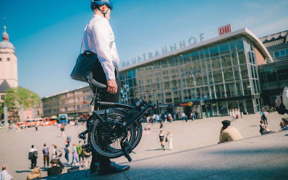 Mann steht mit zusammengeklapptem Brompton vor Kölner Hauptbahnhof.